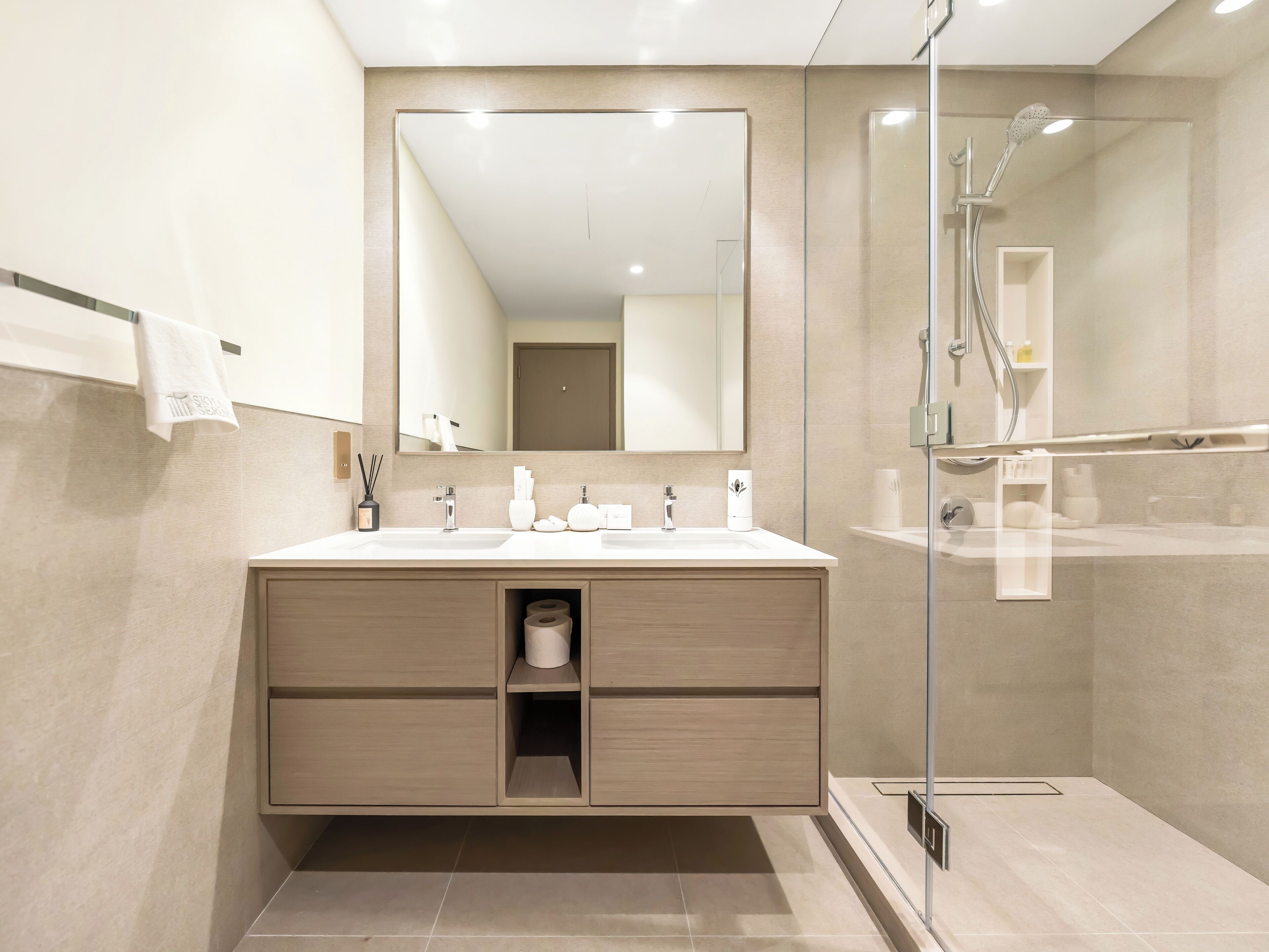 A modern bathroom with a large mirror, two sinks, a shower, and a toilet.