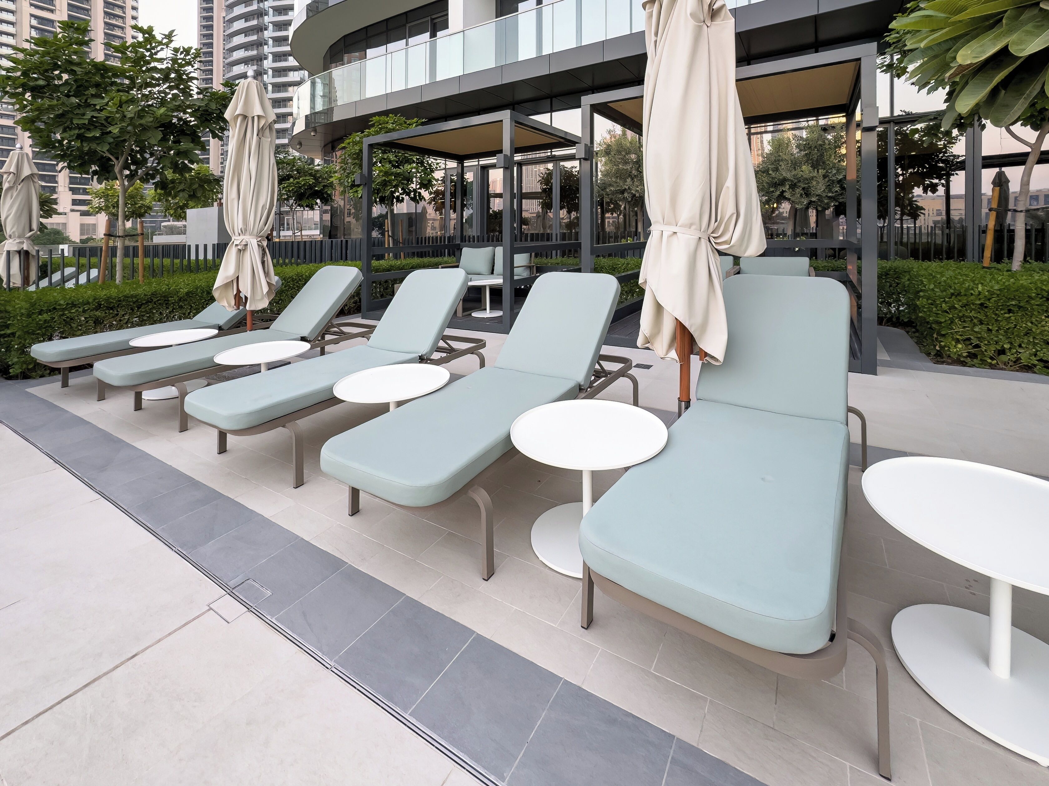 Eine Reihe hellblauer Liegestühle mit weißen Tischen, angeordnet auf einer gefliesten Terrasse, im Hintergrund ein modernes Gebäude.