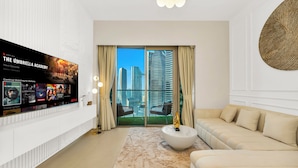 A modern living room with a large TV displaying a series, a sofa, a coffee table, and a view of tall buildings through a glass door.