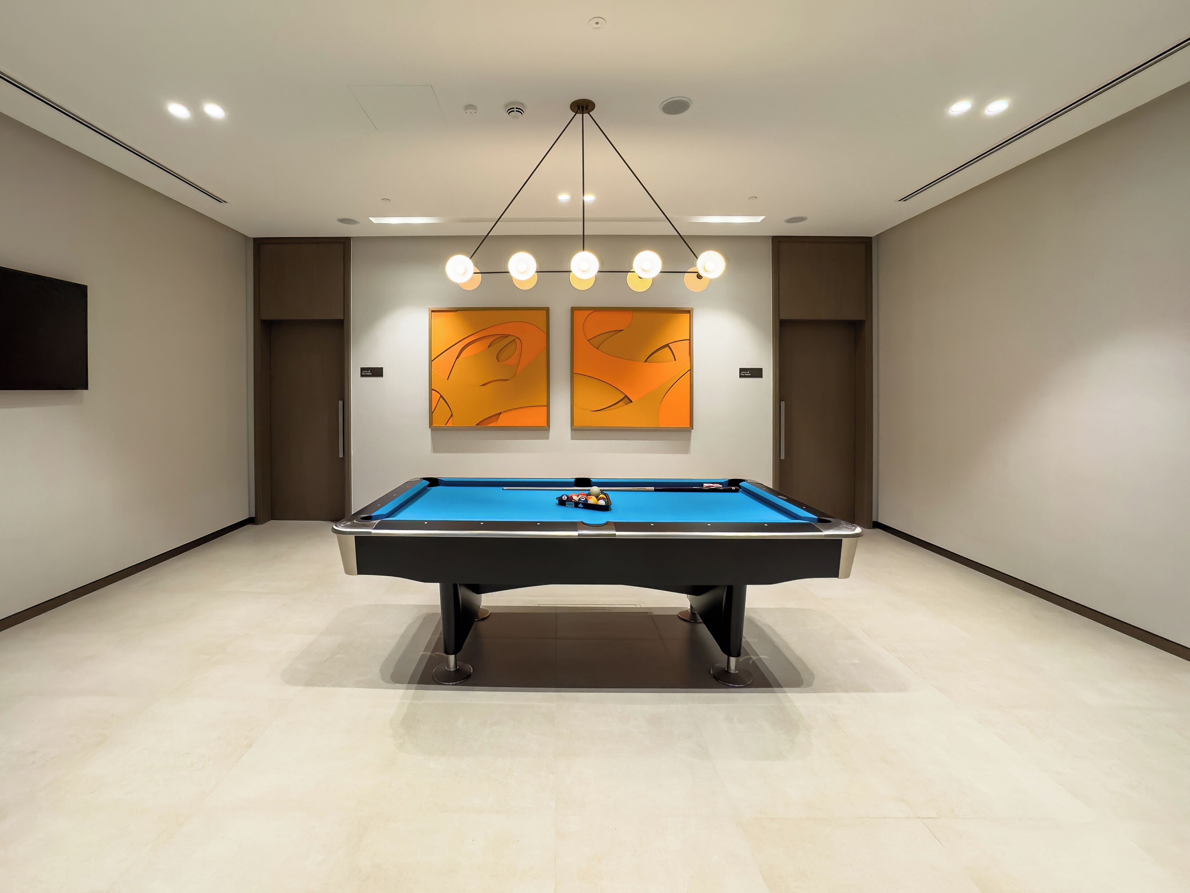 Une table de billard dans une pièce au design moderne, comprenant un luminaire au plafond et deux tableaux abstraits accrochés au mur.