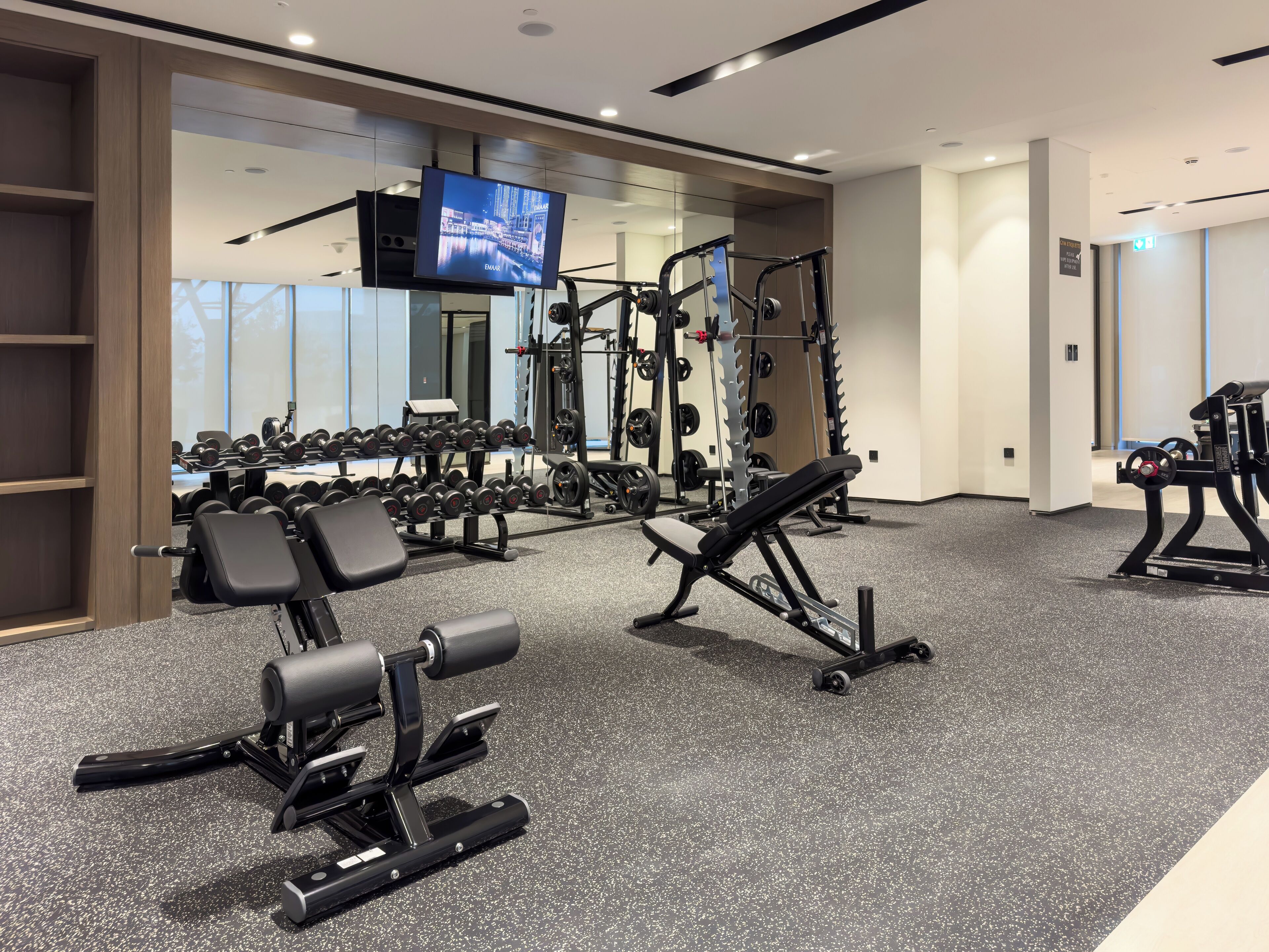 Une salle de sport moderne équipée de divers appareils d’exercice, notamment des haltères, des machines de musculation et un écran de télévision diffusant des images d’un paysage urbain.
