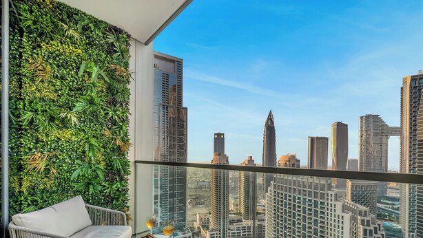 A balcony with a city view, a wicker chair, a small table with a box of chocolates and two glasses of wine, and a green wall with plants.