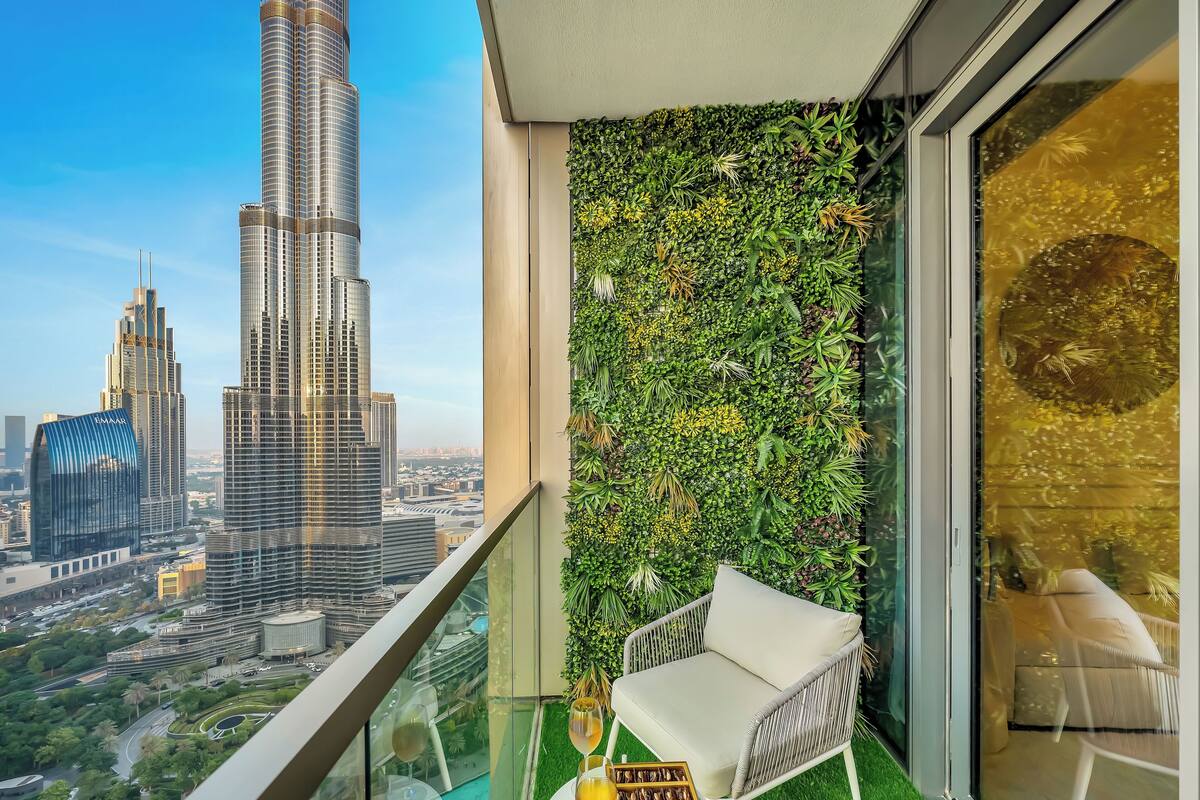 A balcony with a view of the Burj Khalifa and other buildings, a glass railing, a white chair, and a green wall with plants.