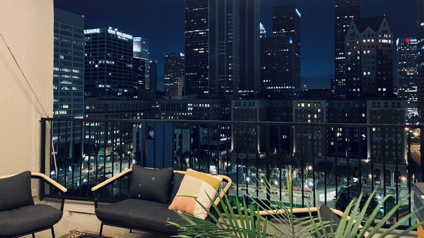 A balcony with string lights, two black chairs, and a view of a city skyline at night. - Sun-Soaked Urban-Chic Retreat in DTLA (Los Angeles)