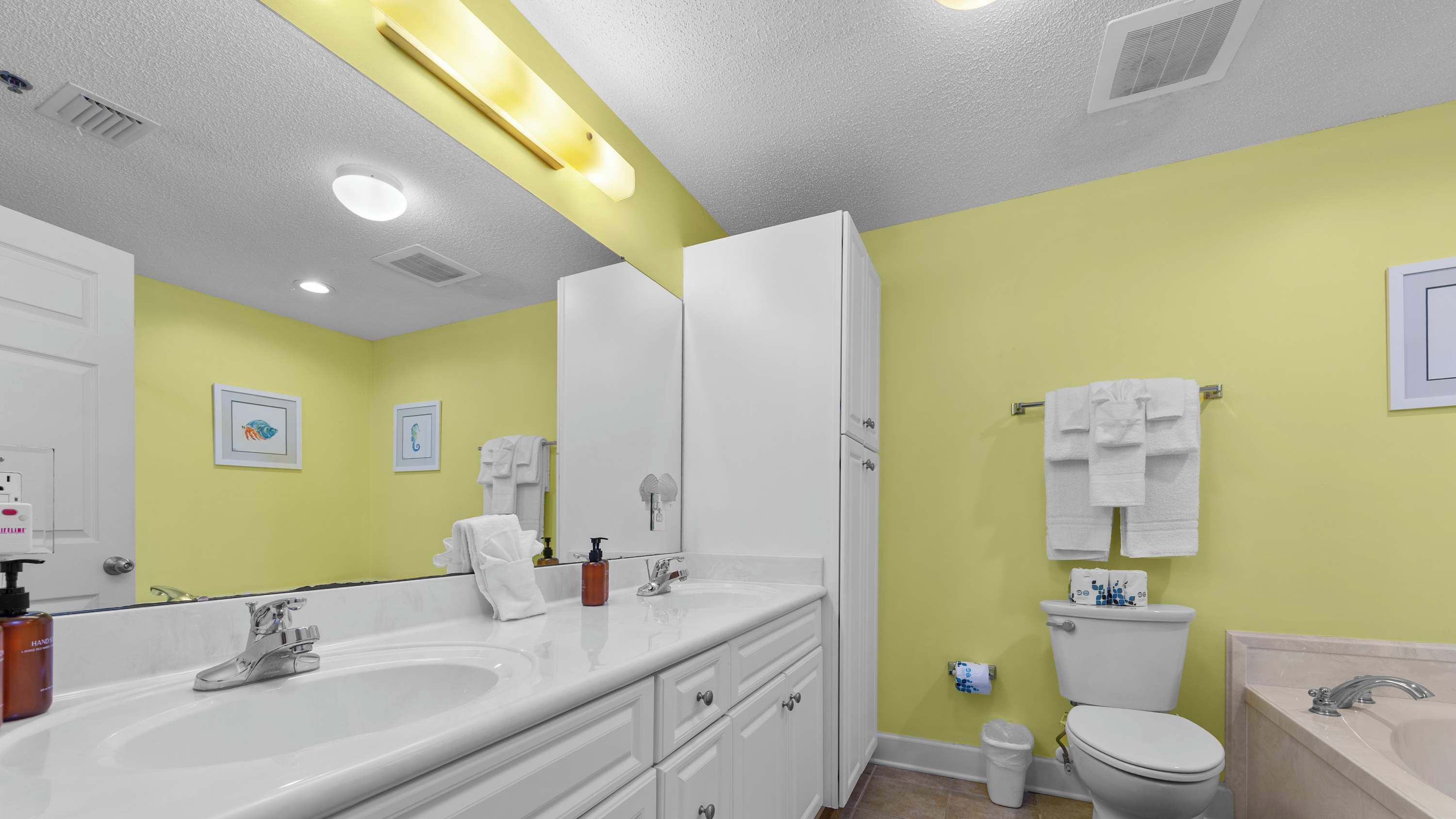 A bathroom with a double sink, a toilet, and a bathtub. There are towels hanging on the wall and a trash can near the toilet.