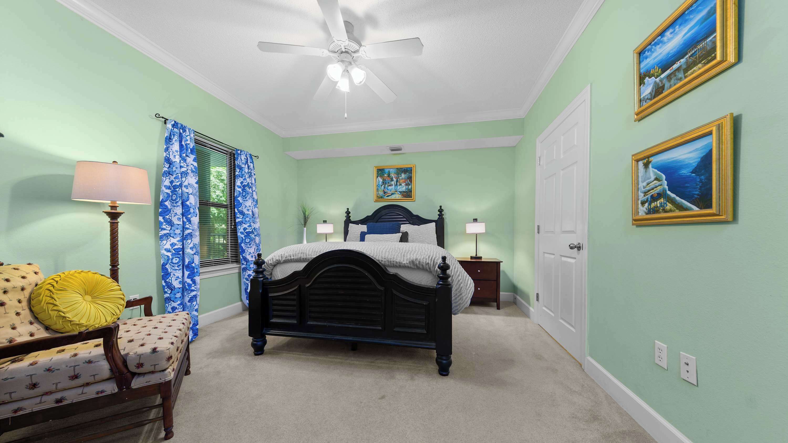 A bedroom with a bed, a chair, a nightstand, a lamp, a ceiling fan, and two paintings on the wall.