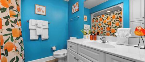 Un baño con paredes azules, un inodoro blanco, un lavabo con espejo y una cortina de ducha con diseño naranja.
