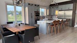 A modern kitchen with grey cabinets, a central island with bar stools, and a large window offering a view of greenery. - Muskoka 5BR Lakefront Cottage · Doe Lake · Sleeps 12+ · Pet-friendly · 4 Season (Gravenhurst)
