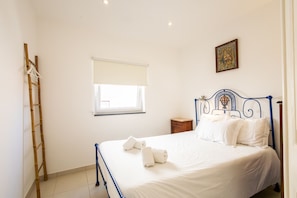 Un lit bien fait, avec du linge de lit et des serviettes blanches, une échelle en bois appuyée contre le mur, une petite table de chevet en bois et une fenêtre avec un store blanc.