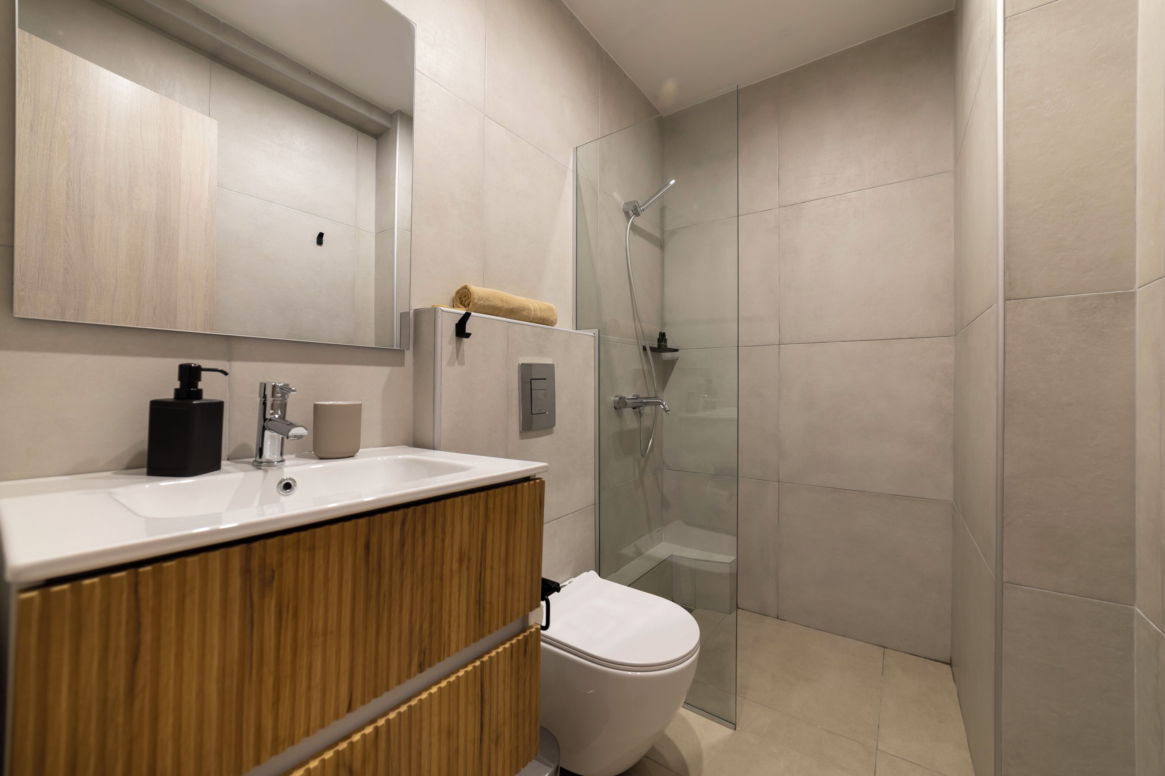 Un baño moderno con un mueble tocador de madera, un lavabo blanco, un inodoro y una ducha con mampara de vidrio.
