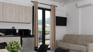 A modern living room with a sofa, a dining table with chairs, a small potted plant, and a kitchen area with a coffee machine. - NS Apartments Nydri (Lefkada)
