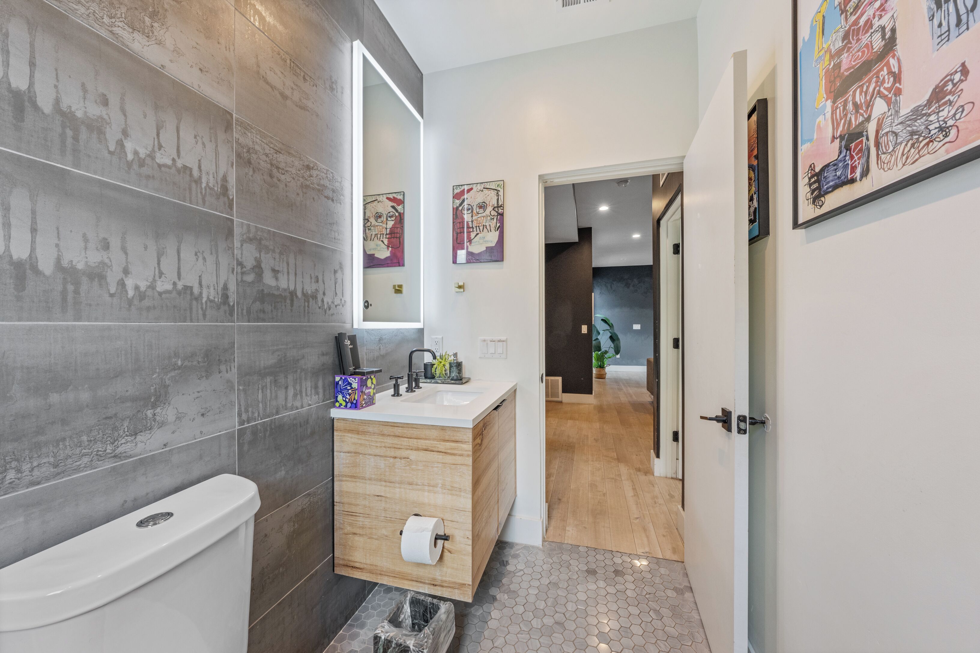 A modern bathroom with a toilet, sink, and mirror. There are two framed artworks on the wall and a hallway leading to another room.
