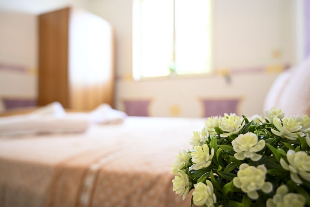 A hotel room with a neatly made bed, a window with curtains, and a bouquet of white flowers on the bed.