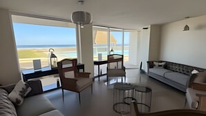 A spacious living area with a large window offering a view of the beach and ocean. There is a sofa, a dining table with chairs, and a small side table. - Lagoon beach white 311 (La Serena)