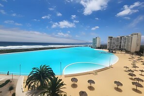 A large swimming pool with a view of the ocean, surrounded by beach umbrellas and palm trees.