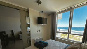 Ein Hotelzimmer mit Bett, einem an der Wand befestigten Fernseher, einem Fenster mit Meerblick und einem kleinen Tisch mit Stühlen.