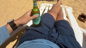 Eine Person entspannt sich auf einem Liegestuhl am Strand, mit einer Bierflasche, einem Sonnenschirm und einem Paar Sandalen.