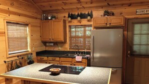 A compact kitchen with wooden cabinets, a stainless steel refrigerator, a built-in island with a kettle, and a window with blinds.