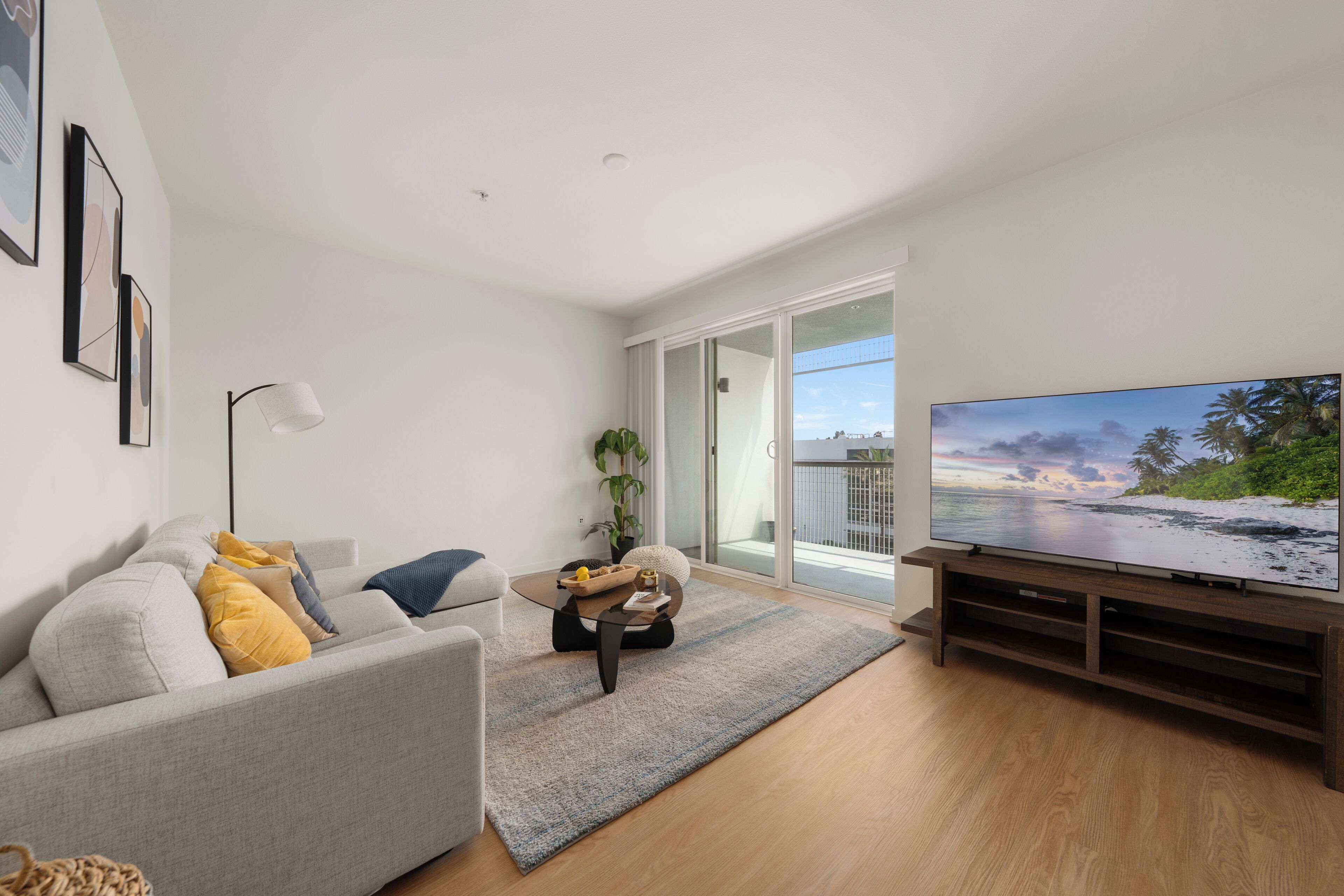 A modern living room with a grey sofa, a coffee table, a flat-screen TV, and a large window with a view of the beach.