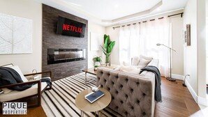 A modern living room with a fireplace, a sofa, a coffee table, and a large plant. - Mayfield Manor-Hot Tub-KingBed-FireTable-BBQ-Wifi (Edmonton)