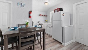 Cocina compacta con alacenas blancas, un refrigerador blanco y un microondas encima. Hay una mesa de comedor con sillas y un jarrón con flores.
