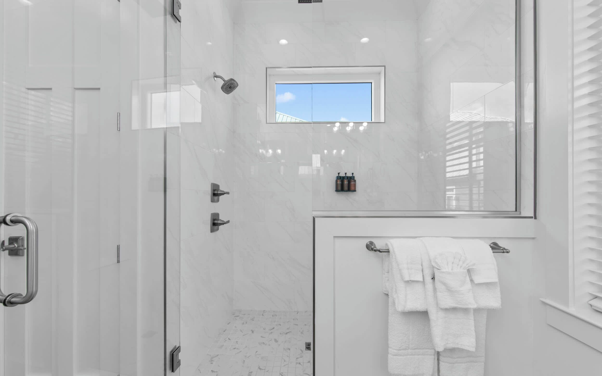 Un espacio de ducha moderno con puerta de cristal, una ventana pequeña y un perchero con toallas blancas.