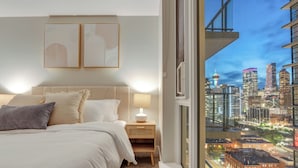 A modern bedroom with a city view, a bed, bedside table, and artwork on the wall. - The Astoria | Steps to Stampede+Saddledome+BMO Center| Free Parking (Calgary)