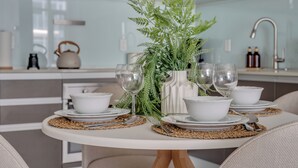 A modern kitchen with a dining table set for four, featuring white plates, bowls, and glassware, accompanied by a decorative plant and a kettle. - The Astoria | Steps to Stampede+Saddledome+BMO Center| Free Parking (Calgary)