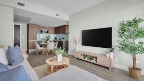 A modern living room with a sofa, a coffee table, and a TV. There is a dining area with chairs and a table, and a kitchen in the background. - The Astoria | Steps to Stampede+Saddledome+BMO Center| Free Parking (Calgary)