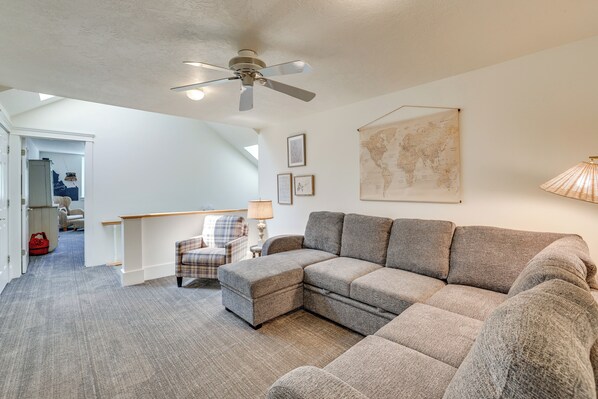 Ein Wohnzimmer mit einem Ecksofa, einem karierten Sessel, einem kleinen Tisch mit Lampe und einem Deckenventilator.