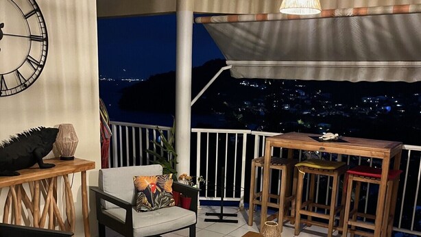 A cozy outdoor seating area with a wooden table, chairs, and a large clock on the wall. - Appart Anse à l'Ane, Les Trois Ilets, with panoramic view 300 m from the sea (LES TROIS ILETS)