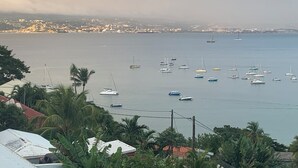 A coastal view with boats, palm trees, and a distant cityscape. - Appart Anse à l'Ane, Les Trois Ilets, with panoramic view 300 m from the sea (LES TROIS ILETS)