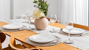 Ein Esstisch ist mit weißen Tellern, Besteck und Weingläsern gedeckt, in der Mitte steht eine Vase mit weißen Blumen.