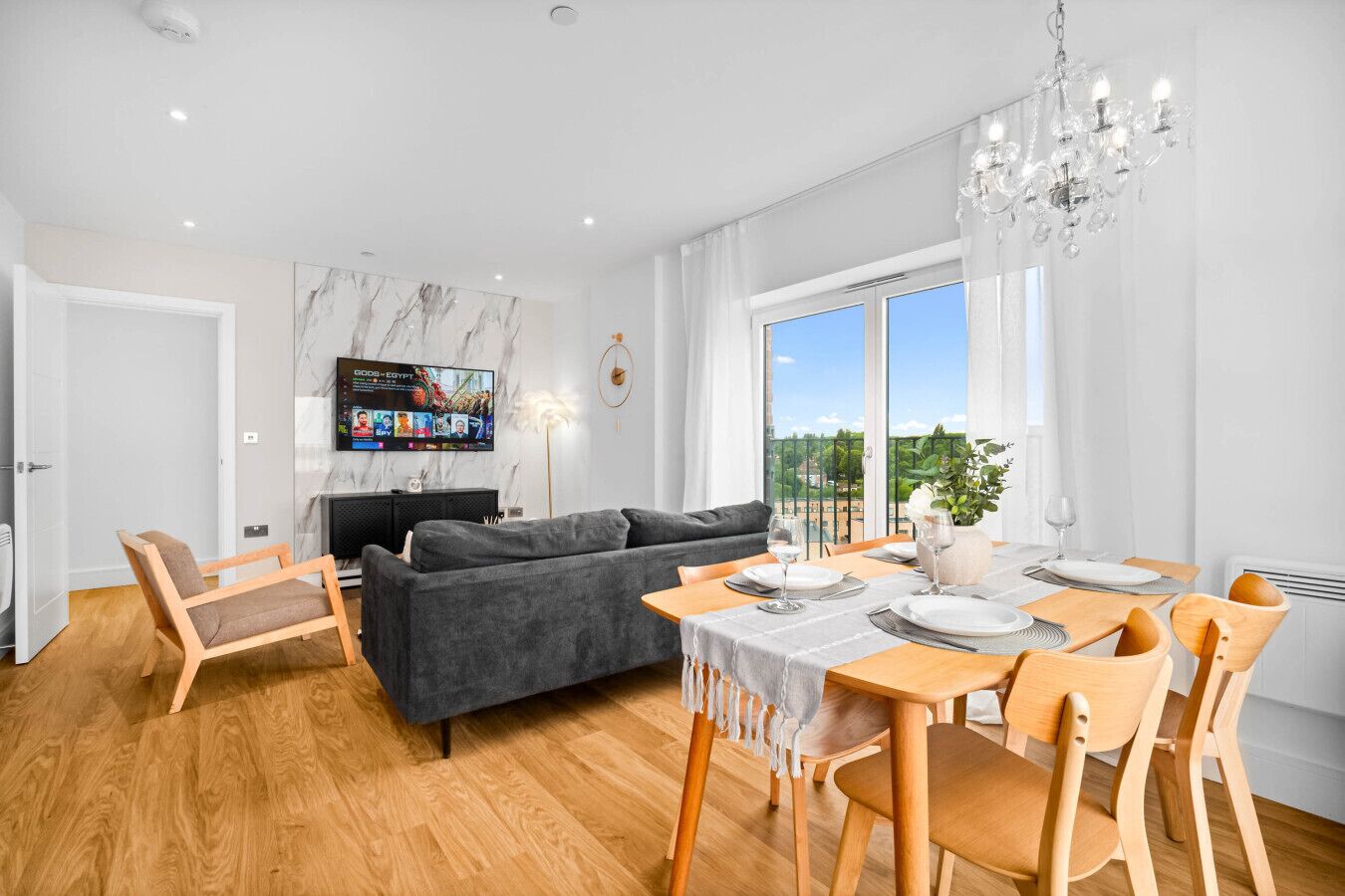A modern living room with a grey sofa, wooden chairs, a dining table set for two, and a TV mounted on the wall.