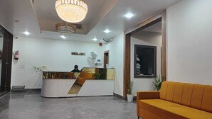A reception area with a reception desk, a chandelier, a reception sign, a sofa, and a window. - Hotel grand Lucent  (Mahaban)