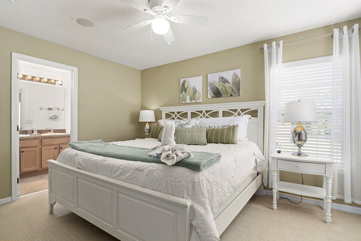 Une chambre à coucher avec un lit blanc, une table de chevet avec une lampe, un ventilateur de plafond et une fenêtre avec des stores.