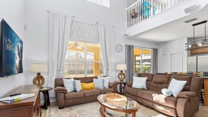 A modern living room with two brown sofas, a glass coffee table, and a large window with curtains. - Luxury 2850sf Rental | Water + Conservation Views | Near Clubhouse | Gated (Davenport)