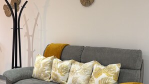 A grey sofa with floral patterned cushions and a mustard throw blanket, positioned against a white wall with decorative wall art.