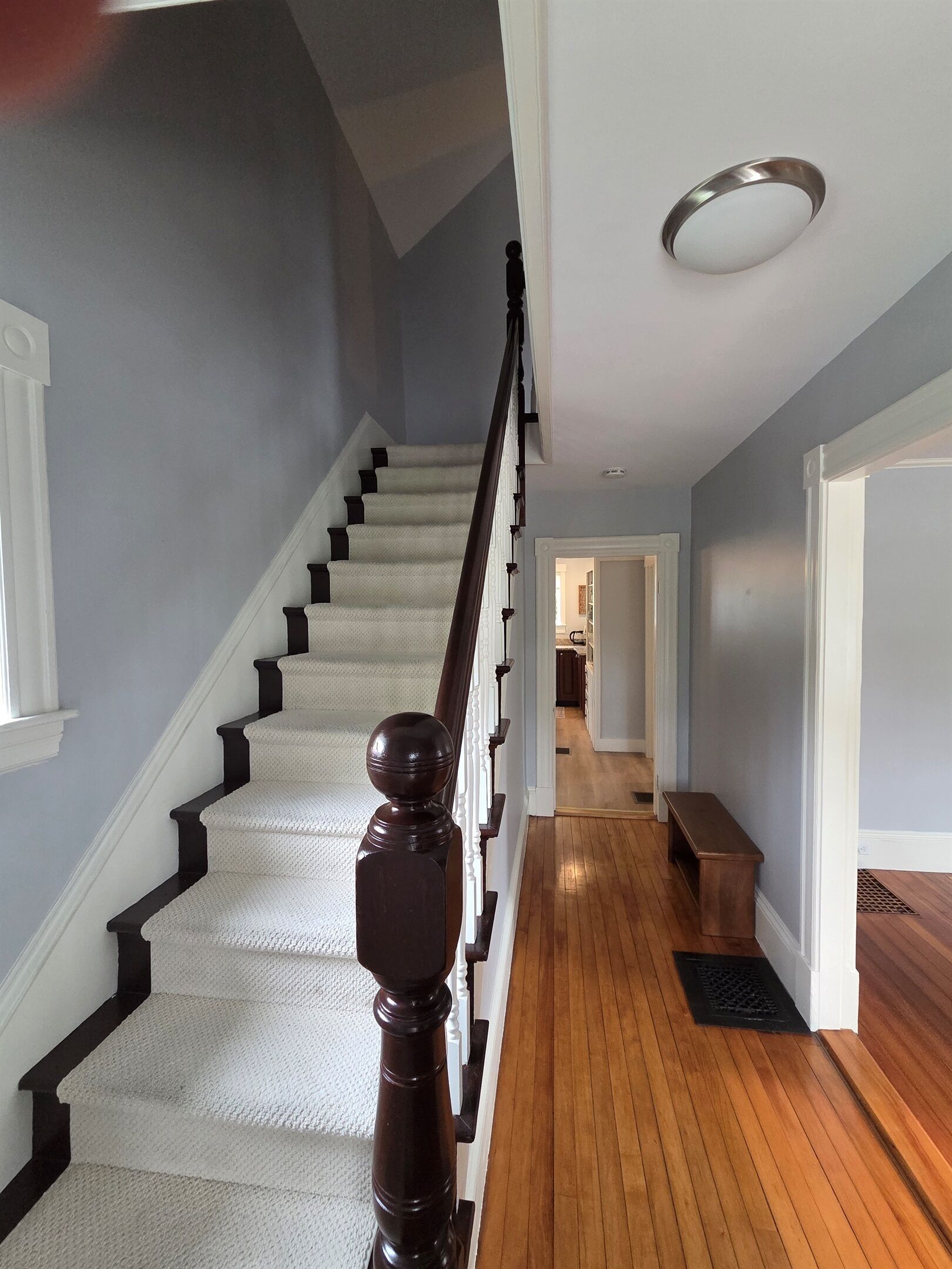 A staircase with a wooden handrail and white carpeted steps leading to a hallway with wooden flooring and a bench.