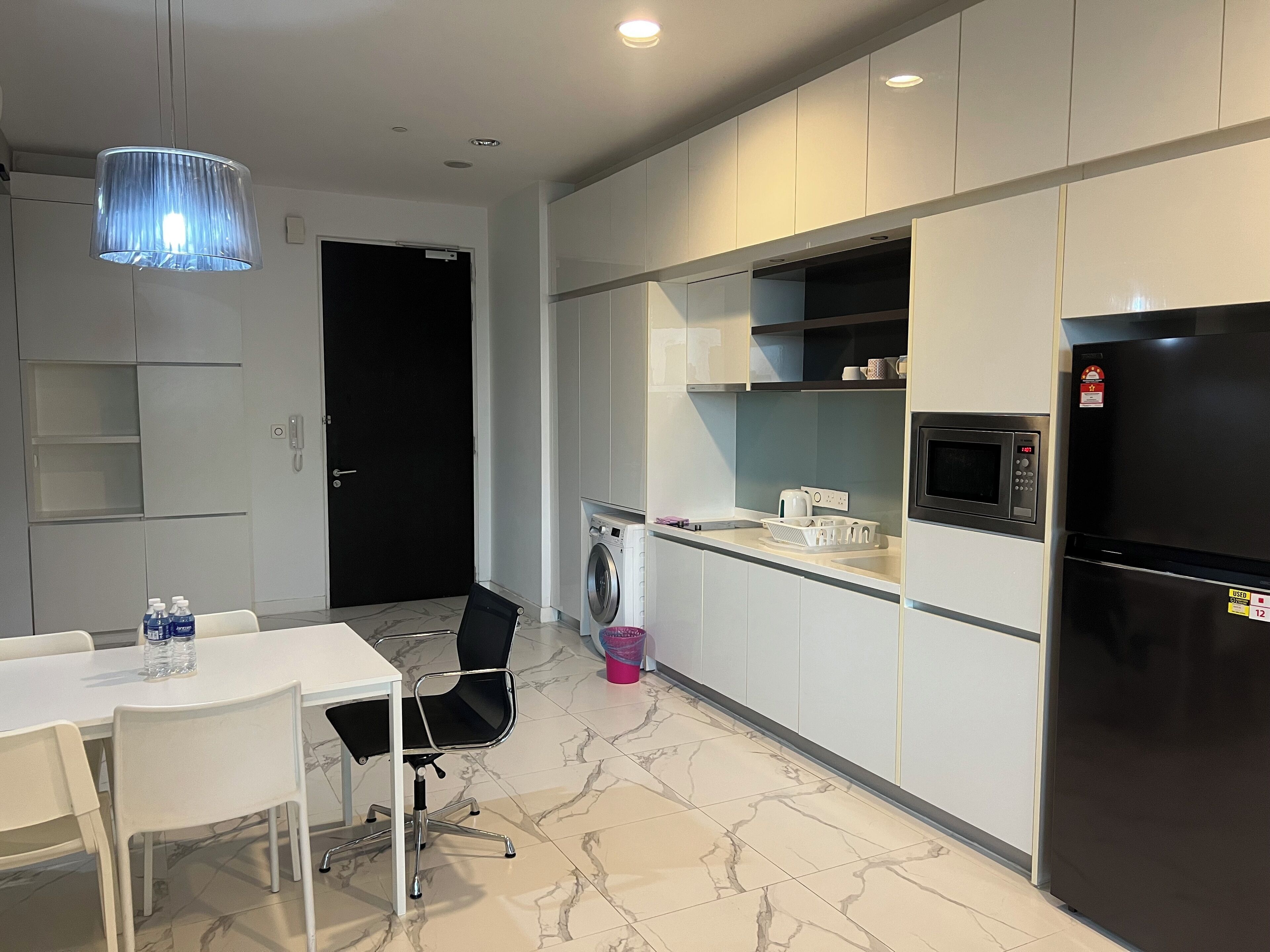 Une cuisine moderne avec des meubles blancs, un réfrigérateur noir, un four à micro-ondes et un lave-linge. On y trouve une table à manger avec des chaises et une suspension bleue.