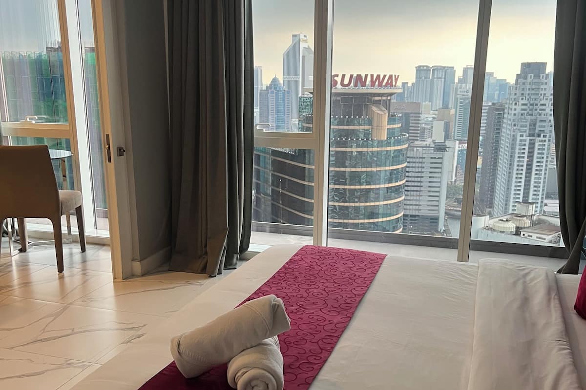 Habitación de hotel con una cama grande, un corredor decorativo rojo, una toalla enrollada y vistas de un paisaje urbano con edificios altos.