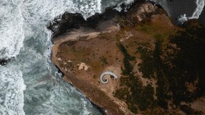 Eine Vogelperspektive zeigt eine Küstenlandschaft mit einer spiralförmigen Struktur in der Nähe des Strandes, umgeben von Vegetation und felsigem Gelände.