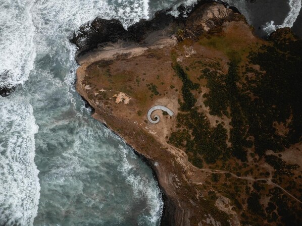 Vue aérienne d’une zone côtière avec une structure en forme de spirale près du rivage, entourée de végétation et d’un terrain rocheux.