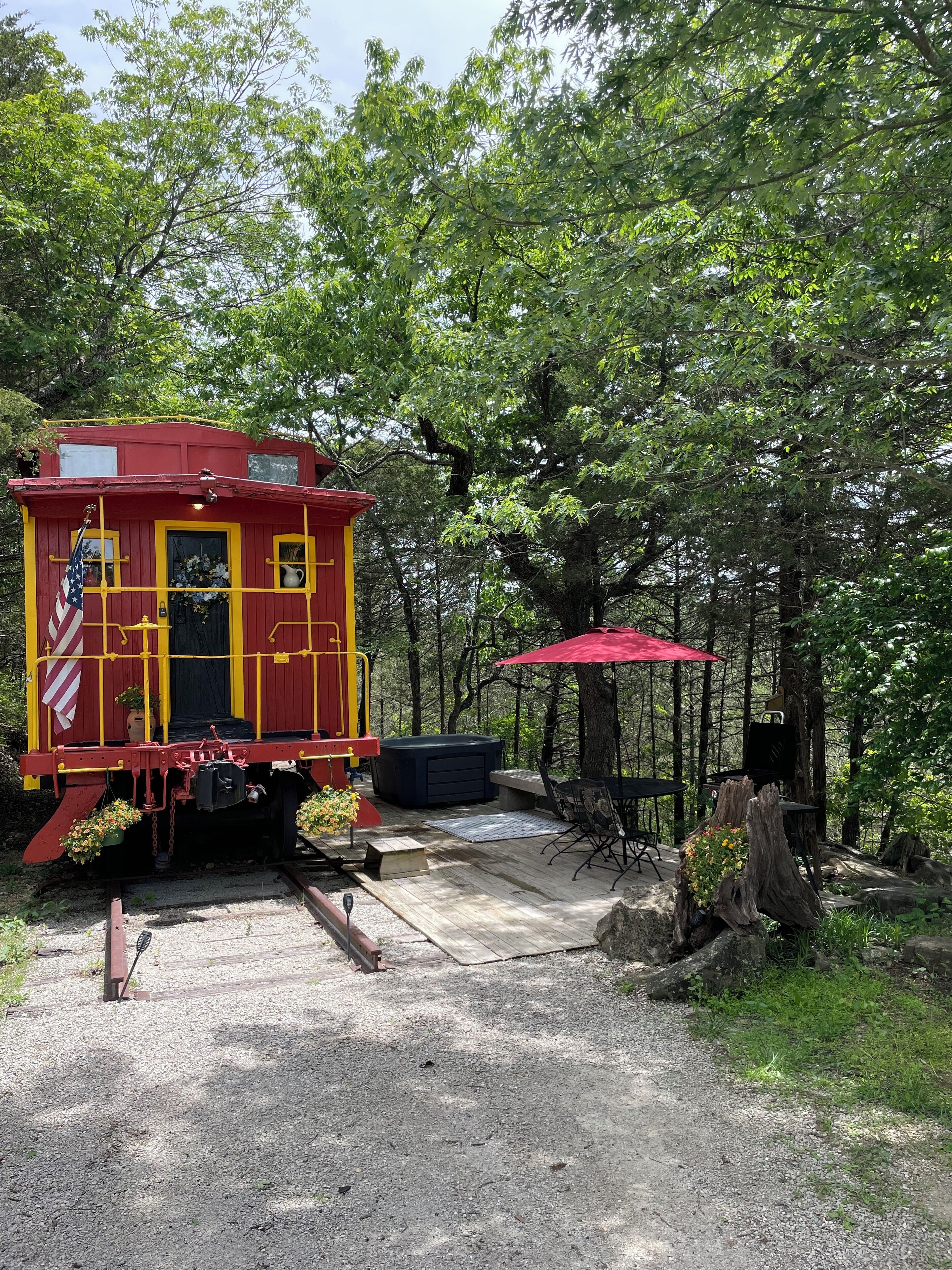 A red caboose with an American flag, a black grill, and a red umbrella in a wooded area.