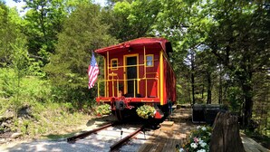 Un vagón de tren rojo con bandera estadounidense, situado en un sendero de grava con tablones de madera, rodeado de follaje y una estructura de madera.