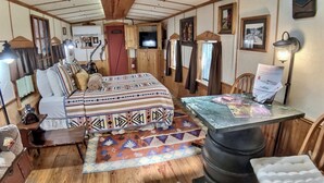 Interior acogedor de una cabaña con una cama, una mesa con papeles, una lámpara y un barril grande.