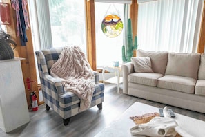 Ein Wohnzimmer mit einem Karo-Sessel, einem beigen Sofa, einem kleinen weißen Tisch und einem hängenden Buntglasfenster.