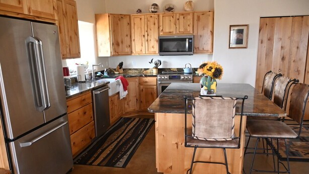 A kitchen with wooden cabinets, a stainless steel refrigerator, a microwave, and a dining area with a marble countertop and bar stools. - Unique top of the world straw bale cottage has ASTOUNDING views. (Catalina)