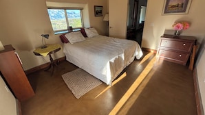 A bedroom with a bed, a desk, a chair, a dresser, a window with a view, and a door leading to another room. - Unique top of the world straw bale cottage has ASTOUNDING views. (Catalina)