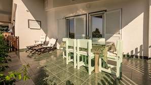 Un patio con mesa y sillas, cercado por una valla de madera y con una pared blanca con contraventanas.